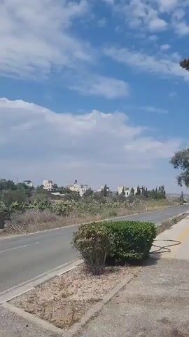 The video captures a roadside scene under a partly cloudy sky. A paved road runs horizontally across the frame, with a gravel shoulder and a small landscaped area in the foreground. Beyond the road, there's a dense patch of prickly pear cacti and other vegetation. In the middle ground, several modern-looking white buildings with flat roofs are visible, nestled among trees and further vegetation. The overall impression is of a somewhat arid, rural or suburban landscape.
