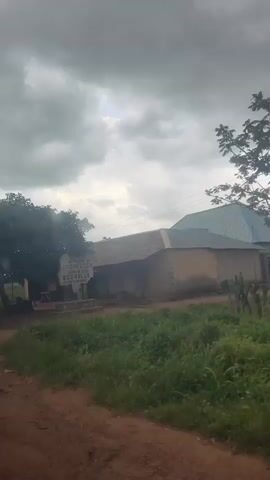 The video begins showing a sign with the text WELCOME TO COGIN LCCU. The video pans to the right, showing a tree and a building. Then it shows a dusty road and some greenery. The video continues to pan to the right showing a red bucket on the side of the road. It then pans to show a building with a metal roof and trees.