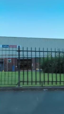 The video shows a view from inside a vehicle, likely a car, moving past an industrial area. The foreground shows the interior of the car, including a side mirror on the right. A black metal fence runs horizontally across the middle of the frame, separating the road from a grassy area with bushes and trees. Behind the fence, a long, light-colored industrial building is visible, stretching across the background. A sign with the words "Autobase Industrial Park" is seen to the right of the building. There are some objects on the ground near the fence, including a blue plastic item. Several street lights and security cameras are mounted on poles within the industrial park. The sky is a clear, pale blue. There are no people clearly visible in the video.