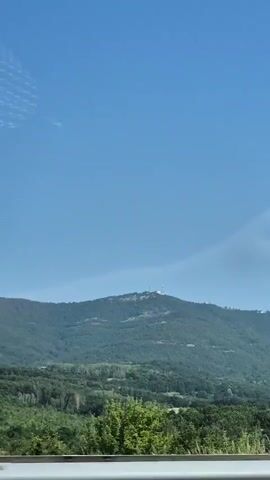 This is a video shot from a moving vehicle. The camera is pointed towards a large hill covered in green trees. There are some scattered buildings and structures visible on top of the hill, along with a tall white tower. The sky is a clear blue with a few wispy clouds. In the foreground, there are glimpses of trees and a white barrier, suggesting the vehicle is on a road. The video is taken during the daytime, with ample sunlight.