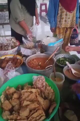 The video shows an outdoor food stall with several people. In the foreground, a woman in a blue hijab and pink shirt is placing food items into plastic bags. She is holding a piece of fried food in her left hand and a plastic bag in her right. In front of her is a large green bowl filled with more fried food. To her right, another person's hand is visible reaching for a spoon in a bowl of sauce. Behind her, there are several large pots and bowls containing food, including a pot of reddish stew, a bowl of chopped green vegetables, and a bowl of white rice. Two other people are visible in the background, one wearing a blue cardigan and a red headscarf, and another in a black headscarf. They appear to be interacting with the food stall. The environment appears to be a paved area with some outdoor seating and vegetation in the background.