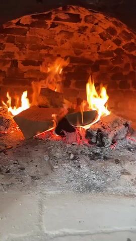 The video shows a fire burning inside a brick oven. Several pieces of wood are actively on fire, with orange flames rising. A pile of ash and charred wood is visible beneath the burning logs. The bricks forming the oven's walls and ceiling are various shades of orange and red. The fire illuminates the interior of the oven, creating a warm glow.