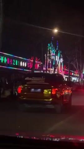 The video shows a nighttime street scene. A black Range Rover drives on the right side of the street.  On the left, a long building is decorated with bright, colorful neon lights. There are several smaller parked vehicles beside the building, and a few pedestrians are walking in front of it.  A street light shines above.  The video is shot from inside a vehicle, looking out the windshield, and some of the dashboard is visible.  Red tail lights from another car are visible in front of the camera vehicle.