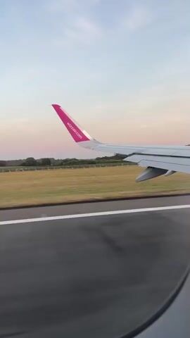 This is a point-of-view shot from an airplane window, looking out at the wing and the runway during takeoff or landing. The airplane's wing, with the Wizz Air logo on its pink wingtip, is prominently in the foreground, partially obscuring the view of the runway. The runway markings are visible, and beyond it, a grassy field stretches out, bordered by trees and a distant treeline. The sky is a pale blue with streaks of pink and orange, indicating either sunrise or sunset. There are no people visible in the frame. The scene captures the motion of the plane as it either accelerates down the runway or decelerates after landing, with the landscape blurring slightly due to the movement.