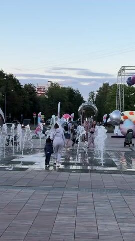 A group of people, including children and adults, are gathered in an outdoor public space featuring a water fountain. The ground is paved with square tiles. Several large inflatable sculptures, some resembling cartoon characters, are visible in the background, along with trees, buildings, and a stage structure. The sky is overcast with clouds. The water from the fountain is spraying upwards, creating splashes around the people. Some individuals are walking through the water. The lighting suggests it is either early morning or late afternoon.