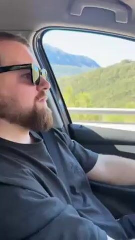 The video shows the interior of a car driving on a highway. A man with a beard and sunglasses is seen riding in the passenger seat and then driving. He is wearing a dark shirt and a gold watch. The car's dashboard is visible, with a phone mounted on a holder. The highway runs alongside a mountain range covered in green trees under a clear blue sky. The video is shot from within the car. The driver appears to be enjoying the drive.