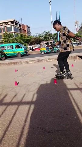 A medium shot shows two people rollerblading between cones on a paved area near a busy street. The primary focus is on the individual closest to the camera, wearing a patterned shirt and black pants. This person maneuvers around pink cones placed in a line. In the background, another person in a white shirt and black pants also skates between the cones. The location seems to be an urban area, indicated by buildings, traffic, and streetlights. The camera is positioned behind a metal fence, casting long shadows on the ground.