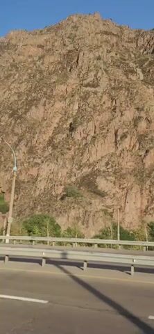 The video shows a view from a vehicle traveling along a highway. On the right side of the highway, there is a steep, rocky mountain face with sparse vegetation at its base. The highway has multiple lanes and a guardrail separates it from the mountainside. Utility poles and wires are visible along the side of the road, and a few buildings are situated near the mountains. The sky is clear and blue. The camera pans slightly to the right as the vehicle moves forward, capturing the mountainous landscape and the roadside infrastructure. There are no people clearly visible in the video.