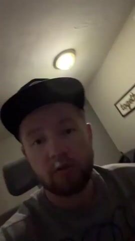 A man wearing a black baseball cap and a grey t-shirt with a black design sits in a chair and rests his head on his hand. He is looking towards the camera. The room is dimly lit, with a light fixture visible on the ceiling and a framed picture on the wall that says "together".