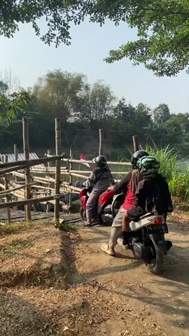 Two motorcycles are parked on a dirt path. One motorcycle is red and the other is white. Three people are on or near the motorcycles. The red motorcycle is ridden by a person wearing a red helmet, a patterned shirt, and dark pants. A second person wearing a helmet and a black jacket is a passenger on the white motorcycle. The white motorcycle is ridden by a person wearing a helmet, a red shirt, and dark pants. The motorcycles are positioned facing a wooden structure. The wooden structure is made of bamboo and appears to be a pier or walkway over water. Trees and foliage are visible in the background. The sky is clear and bright. The lighting suggests it is daytime.