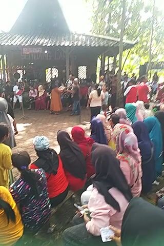 Here is a description of the video.

The video shows a large group of people kneeling on the ground. They are gathered outside a structure with a sign that says "SENDANG LARAS." There are trees in the background and sunlight is visible, indicating that it is daytime. The people kneeling are wearing a variety of clothing. Some are wearing head coverings. There are also people standing and sitting in the background. It appears that the people are observing some kind of solemn ritual.