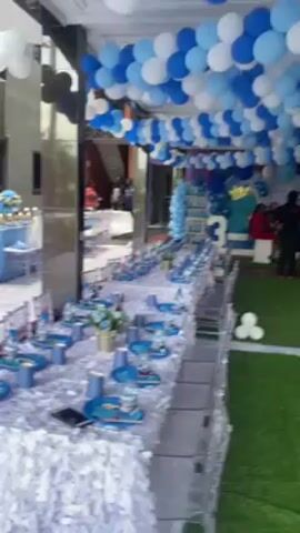 The video shows an event space decorated for a child's birthday party. There are several long tables covered with patterned white tablecloths, set with blue cups, plates, water bottles, and small flower arrangements. The tables are flanked by transparent chairs. The ceiling is adorned with a garland of blue and white balloons. At the end of the tables, there is a decorated area with a large number "3," likely indicating the child's age. Several people can be seen near this area, including adults wearing red and black shirts. The floor is covered with green artificial turf. The space appears to be partially outdoors, possibly under a covered patio or tent-like structure.