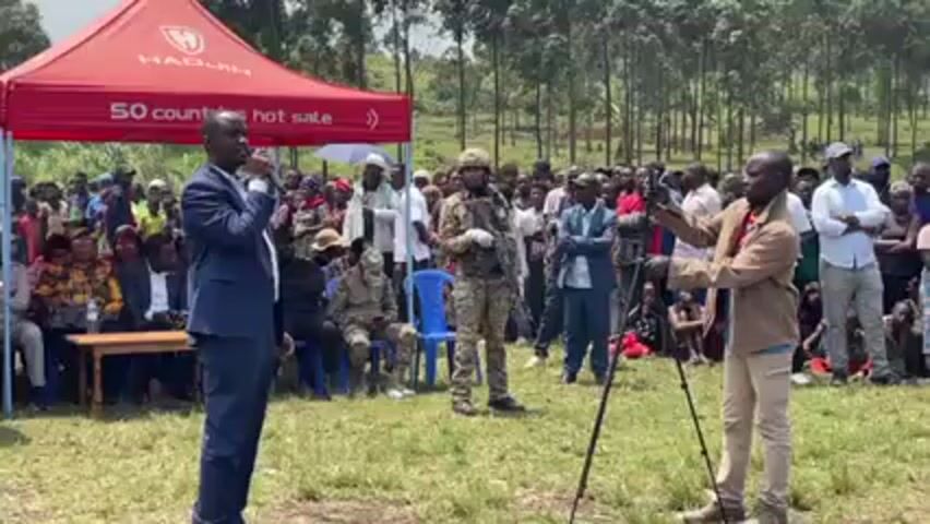 A man in a blue suit is speaking into a microphone in front of a crowd of people, a videographer, and an armed soldier. He is standing on a patch of grass, with several people seated at a table behind him. Two red canopies, with the words "HAQUIN" and "50 countries hot sale" printed on them, are behind the table. Behind the crowd is a forest. The videographer is pointing his camera at the man in the suit. The armed soldier is standing at attention.