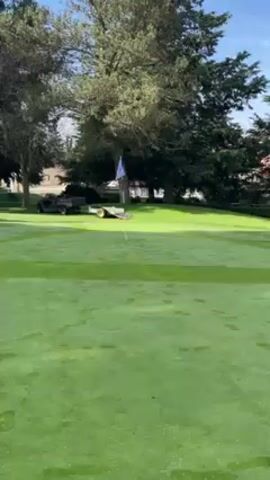 The video shows a man mowing a golf course green. The man is wearing a yellow hard hat and dark clothing. He is walking behind a red and silver lawn mower, moving from right to left across the screen. A golf flag is visible on the green, along with a golf cart and trailer in the background. The environment is lush and green, with trees surrounding the green. The sky is blue and partly cloudy.