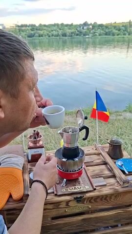 A man is drinking coffee from a white cup by a lake. In front of him is a wooden table with a Moka pot on a stove, a coffee grinder, a glove, and a Romanian flag. The lake has a calm surface reflecting the sky. The background shows trees and a distant building. The scene is captured outdoors during the day, possibly at sunset, given the lighting.