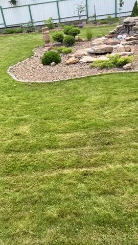 A drone shot pans across a manicured backyard. The yard features a lush green lawn that has recently been mowed, with visible lines from the mower. Beyond the lawn, a landscaped area showcases a rock garden with a small waterfall cascading over stones. Various shrubs and plants are strategically placed around the rock garden, which is surrounded by decorative gravel. A white picket fence encloses the property in the background, and trees are visible beyond the fence. The sky is overcast.