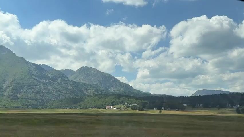 The video shows a wide shot of a mountainous landscape with a cloudy sky. In the foreground, there is a dry grassy field. In the middle ground, there is a green forested area with a few scattered buildings, suggesting a small village. Behind the trees, rugged mountains rise, with some appearing to be covered in vegetation and others showing rocky faces. The sky is blue with large, fluffy white clouds. The camera appears to be moving slightly, giving a sense of travel. A blurry blonde person's hair is visible on the right side of the frame, indicating the video is likely taken from inside a vehicle.