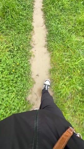 The video shows a first-person view of someone walking along a dirt path. The person is wearing black pants, a black jacket, and black and white sneakers. The path is narrow and surrounded by green grass. There are some yellow flowers scattered in the grass. The camera is shaky, following the motion of the person walking.