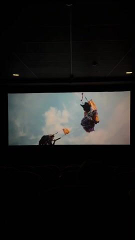 The video shows a movie being viewed in a dark theater. The movie depicts a young man looking urgently at something, possibly holding a tool or weapon. Another scene shows a woman looking up, seemingly trapped or tangled in a parachute or some other material. A wide shot shows a landscape with a river or body of water running through it. The last scene shows someone on the ground, possibly injured, with a colorful parachute or similar object lying nearby. The environment appears to be grassy, possibly mountainous.