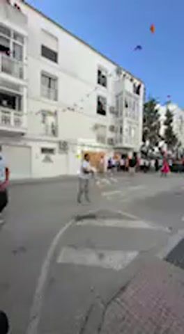 The video displays a vibrant street scene in Vejer de la Frontera, Spain, under a bright blue sky. Whitewashed buildings with balconies line the street, adorned with strings of colorful flags or bunting, suggesting a festive event. Several kites are visible flying high in the distance.

Initially, a person wearing a light-colored shirt and dark trousers stands in the middle of the street, facing away from the camera. Further down the street, a crowd of people is gathered, though their details are blurred. The camera then pans downwards and to the right, revealing the sidewalk, a metal manhole cover, and green foliage along the base of the building on the right. In the final moments of the video, a pair of feet wearing sneakers comes into view, indicating the camera operator has either sat down or lowered the camera. The architectural style and clear weather are characteristic of the region. No explicit text is discernible.
