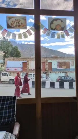 The video is filmed through a wooden-framed window, looking out onto a street. Prayer flags are strung across the top of the window, with pictures of food hanging above them. The street scene includes buildings, vehicles, and two individuals dressed in maroon robes and red headscarves, walking away from the camera. One individual appears to be speaking on a mobile phone. The environment suggests a mountainous region under a cloudy sky.