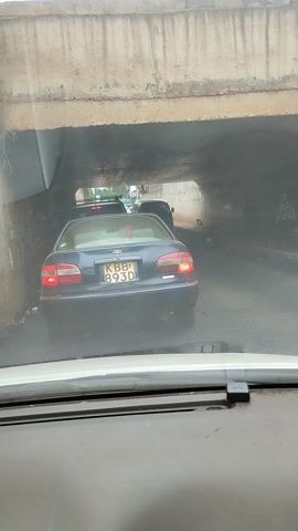 The video shows a view from inside a vehicle looking out its side window, capturing a scene of traffic in an underpass or tunnel. On the left, concrete walls of the structure are visible. Ahead, a blue Toyota sedan with license plate "KBB 893D" is stopped or moving slowly. To its left, a white van with license plate "KBJ 550B" is also visible. Other vehicles are further down the tunnel, which appears to be dimly lit but indicates daytime outside. The overall scene suggests traffic congestion within an underpass in an urban or semi-urban area.