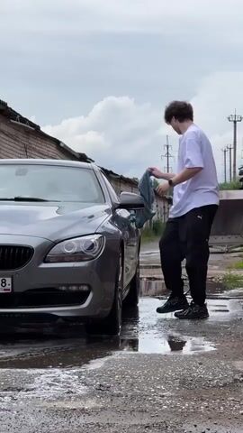 A person is cleaning a car in a car wash. The car is a grey BMW, parked on a wet, muddy surface. The person, wearing a white t-shirt and black pants, is holding a blue cloth. The setting appears to be an outdoor car wash, with brick garages in the background. The sky is cloudy, suggesting it might be an overcast day. The ground is covered in puddles and dirt. The license plate of the car is visible and reads "X677YY 18 RUS". The overall mood is one of routine maintenance or a car wash in progress.