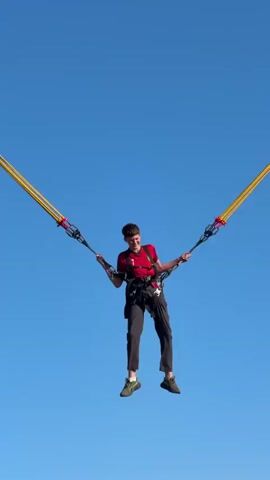 A young person wearing a red shirt, black pants, and a harness is suspended in the air against a clear blue sky. They are connected to two yellow elastic cords extending upwards and outwards. The person's arms are outstretched, holding onto the cords, and they appear to be smiling. The background is a vast expanse of blue sky, with no other discernible objects or structures visible except for a small, white object at the very bottom of the frame. The overall impression is of a recreational activity involving being launched or suspended in the air.