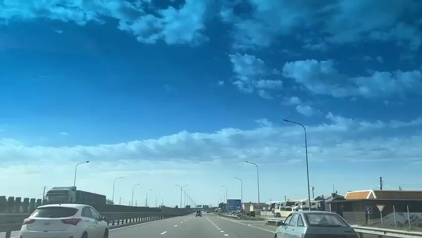 The video shows a driver's view of a highway on a sunny day with a partially cloudy blue sky. The highway is lined with streetlights and has multiple lanes. There are several cars driving on the highway. On the right side of the road, there are some buildings and parked trucks.  A sign indicating a nearby location is also visible.