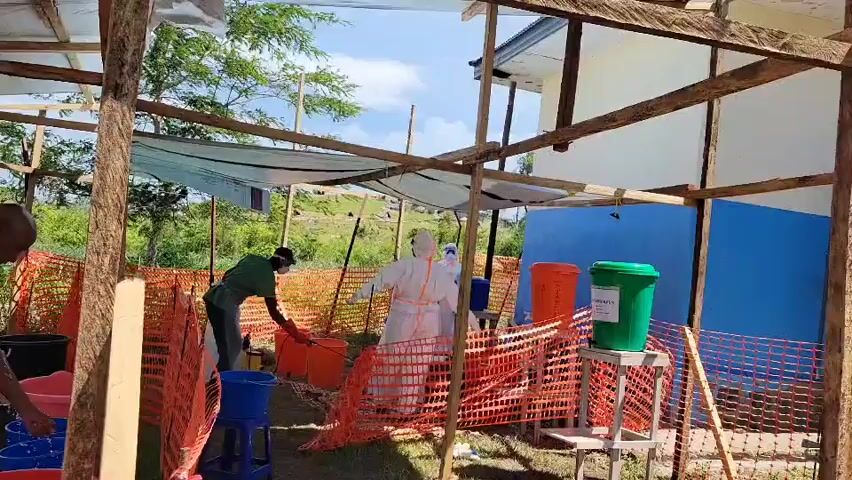 This media file captures an outdoor health or sanitation station in Tchere, Democratic Republic of the Congo, under a bright, sunny daytime sky. The scene is set beneath a makeshift shelter constructed from wooden poles and a white tarpaulin, offering shade to the operations below.

In the center of the frame, two individuals are prominently visible, dressed in full white personal protective equipment (PPE) suits, including hooded coveralls. One of them is holding a spray device, seemingly engaged in a process of disinfection or decontamination. Another person in a green shirt and dark pants is positioned to the left, bending over several blue buckets, suggesting involvement in preparing or managing liquids.

The area is organized with orange safety netting acting as barriers, defining specific zones for activity. Numerous plastic buckets in various colors, including orange, blue, black, and pink, are scattered on the ground, likely serving different purposes such as holding water, cleaning solutions, or waste. Two larger bins are elevated on stands: one orange and one green. The green bin features a white label that clearly reads "INFECTIOUS WASTE," indicating its designated use for hazardous materials.

In the background, a simple building with blue and white walls is visible, flanked by lush green trees and a distant hill. The overall setup, characterized by PPE, disinfection equipment, and segregated waste disposal, strongly suggests a public health protocol or emergency response in a rural or semi-rural setting.