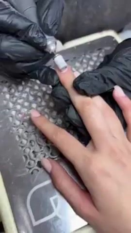 A pair of hands wearing black gloves is visible in the frame, performing a manicure on a hand with light skin tone. The right hand holds the other hand steady, while the left hand uses a rotary tool with a cylindrical attachment to shape or smooth the nail. The hand being manicured rests on a metallic mesh surface, with the fingers slightly spread apart. The nails are painted in a light, neutral color. The backdrop is a dark, out-of-focus surface.