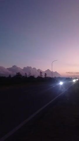 The video captures a serene sunset scene on a highway in Hulawa, Indonesia. The sky is painted in hues of purple and pink, with a few fluffy clouds scattered across the horizon. The sun is setting, casting a warm glow that is reflected on the road. There are a few vehicles on the road, their headlights illuminating the asphalt. The silhouette of trees and utility poles can be seen against the vibrant sky. The overall mood is peaceful and tranquil, evoking a sense of calm and reflection. The time of day is clearly dusk, as indicated by the colors of the sky and the lighting conditions. The location is identifiable as a highway, likely in a rural or semi-urban area, given the presence of trees and open spaces. There is no visible text in the video.