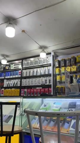 This media file shows the interior of a brightly lit electronics store, likely a mobile phone shop, in Bireuenmeunasahreuleut, Indonesia. The scene is dominated by shelves stacked with mobile phone boxes.

**Subjects:** The primary subjects are the numerous mobile phone boxes and accessories displayed. There are no people visible in the frame.

**Objects:**
*   **Mobile Phone Boxes:** The shelves are filled with white and colored boxes, many of which have branding and model names visible. Examples of text on the boxes include "OPPO A56", "Y11d", "Y05", "C100i", "C85", "C100x", "nubia A36", and "V80".
*   **Accessories:** To the right of the phone boxes, there are racks displaying smaller accessory boxes, likely for earphones and other phone peripherals. Some of these boxes are yellow, green, and black.
*   **Display Cases:** In the foreground, there are glass display cases containing more phone boxes and accessories, also featuring visible text like "POWER SOCK" and "RAPA".
*   **Lighting:** Two prominent LED ceiling lights are visible, illuminating the store.
*   **Furniture:** A partial view of a wooden chair with a white backrest is in the foreground, suggesting a seating area for customers. A stool is also partially visible on the left.

**Scene/Setting:** The setting is a retail environment, specifically an electronics store, characterized by organized shelving and display cases designed to showcase products. The overall impression is that of a busy, well-stocked shop.

**Activity/Event:** The media captures a static view of the store, implying that it is open for business. The arrangement of the products suggests ongoing sales and customer service activities, though none are directly depicted.

**Notable Details:**
*   **Time of Day:** The bright interior lighting suggests it could be daytime, and the store is operational.
*   **Location Cues:** The provided location context of "Bireuenmeunasahreuleut, Indonesia" helps place the store. The types of phone brands and models visible might also be indicative of regional market trends.
*   **Emotions:** No emotions are conveyed as there are no people present. The scene feels neutral and focused on the merchandise.

**Visible Text:** Numerous brand names and model numbers are visible on the product packaging, including "OPPO A56", "Y11d", "Y05", "C100i", "C85", "C100x", "nubia A36", "V80", "POWER SOCK", and "RAPA". Some boxes also feature numerical identifiers like "6" and "12 GB".