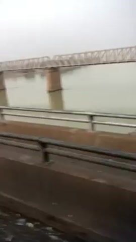 The video depicts a view from a bridge, looking out at another, similar bridge spanning a wide river. The water is a murky, light brown color. Heavy fog or mist obscures the background. The perspective is from a moving vehicle, as indicated by the blurred foreground of the bridge railing. The second bridge is a steel truss structure with supporting piers in the river. The sky is uniformly overcast. The overall scene is gray and muted due to the weather conditions.