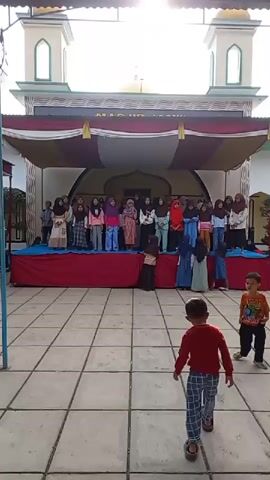 A group of children stand on a stage in front of a mosque, performing. The stage is decorated with red and white fabric. In the foreground, two young boys are running around on a tiled courtyard. The mosque has two minarets and a dome.