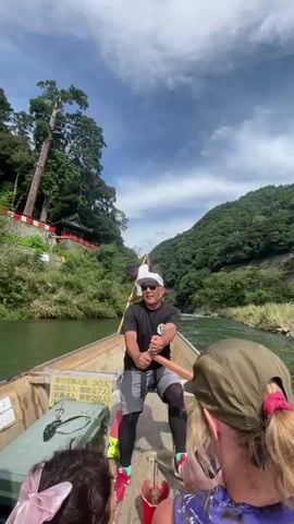 The video shows a group of people in a boat on a river, surrounded by lush green hills and trees. The sky is blue with some white clouds. The boat is wooden and appears to be designed for river excursions. There are at least three people visible. In the foreground, the back of a woman's head wearing a green cap is visible, and next to her, a person with dark curly hair and a pink bow is also visible. In the middle of the boat, a man wearing a black t-shirt, grey shorts, and a white cap is holding a long wooden pole, possibly for steering or propulsion. He is looking towards the camera and smiling. Behind him, another person wearing a white t-shirt and a black hat is also holding a similar wooden pole. They appear to be engaged in a river cruise or tour. In the background, a red-roofed building with a pagoda-like structure is visible on the side of a hill, surrounded by trees. The river flows through a valley, with the water appearing calm and green. There are some Japanese characters written on a sign in the boat.