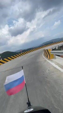 A first-person view from a moving motorcycle on a paved road with a Russian flag attached to the front. The road has a sharp curve with black and yellow striped barriers on the side. In the background, there are mountains and a cloudy sky.  The motorcycle is moving forward.