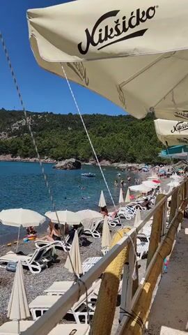 The video shows a crowded beach on a sunny day. The water is clear and blue, with several people swimming and one boat visible. The beach is covered in pebbles, with many lounge chairs and umbrellas set up. People are relaxing on the chairs, sunbathing, and walking along the shore. In the foreground, a wooden railing and part of an umbrella are visible. The background features a lush green hillside sloping down to the sea.