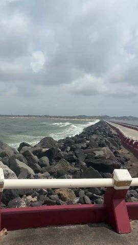 The view is from a pier or walkway, possibly made of concrete, with a red and white railing along the right side. The walkway is bordered on the left by a pile of dark rocks.  Beyond the rocks is a body of water, possibly the ocean, with waves crashing against the rocks. In the distance, a shoreline is visible with a line of buildings and trees. The sky is overcast with gray clouds. The overall scene conveys a sense of being at the edge of the water during a cloudy day.