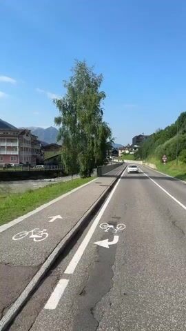 A first-person perspective video shows a road with a separate bike path alongside it. Buildings and trees line the left side of the road. Mountains are visible in the distance under a clear blue sky. Several cars are driving on the road, and some people can be seen walking and cycling on the bike path. The video appears to be filmed in a town or village setting.