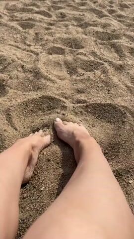 A pair of bare feet are visible on a sandy surface. The feet are partially submerged in the sand, with grains clinging to the toes and ankles. The camera angle is from a first-person perspective, looking down at the feet and legs. The sand is a light brown color and appears to be dry and loose, with some texture and small particles visible. The lighting suggests an outdoor environment, possibly a beach or a sandy area. The legs are fair-skinned, and some faint shadows are cast on the sand. The video consists of a few seconds of still shots, showing the feet in a similar position.