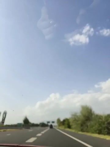 The video shows a view from the inside of a vehicle traveling on a highway. The sky is blue with scattered white clouds. Trees and bushes line the right side of the road, and there are traffic signs and streetlights on the left. A dark-colored car is visible ahead on the highway. The reflection of a person's hands, with manicured fingernails, is visible on the windshield in the upper portion of the frame. The overall impression is a daytime journey on a sunny road.