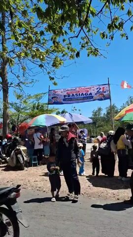 A group of people are gathered outdoors under a bright blue sky, likely for an event. A banner that reads "SELAMAT DATANG PRAJIAGA 21" is visible, suggesting a welcome or celebration. Several individuals are walking and interacting. In the foreground, a man and a young boy are walking together. Behind them, various stalls with colorful umbrellas are set up, with people browsing or working at them. There are also parked motorcycles on the left side of the frame, indicating transportation. The ground appears to be unpaved, with some rocks and dirt. Trees with green leaves provide some shade. The overall atmosphere seems to be lively and festive, with a mix of adults and children present.