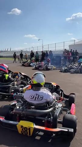A group of go-karts is lined up on a track, engines running and emitting smoke. Each kart has a driver wearing a helmet and racing gear. The karts are numbered, with one prominently displaying "381" and "eifs" on the back. People are standing behind a fence, observing the go-karts. The sky is blue with scattered clouds. A few tents are visible in the background along with flag poles with flags. The ground is gray asphalt. The camera is positioned behind one of the go-karts, looking toward the rest of the group.