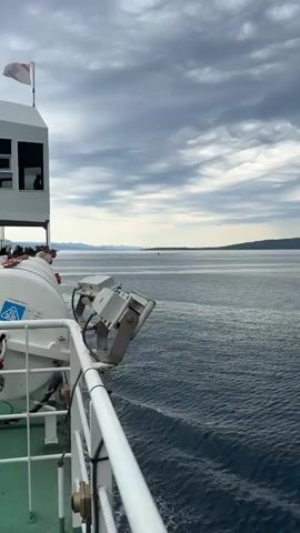 A drone-like view is shown from the deck of a ferry. The water is blue and choppy, with a distant island or coastline on the horizon. The sky is cloudy with patches of blue. On the deck, there is a white railing, a life raft, and some equipment. A few people are visible on the deck.