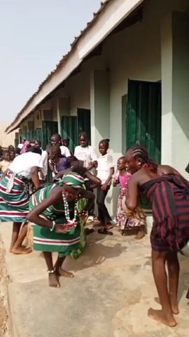 In front of a light-colored building with green windows, two women perform a dance. One is adorned in a green and white striped garment, along with a matching headband and beaded jewelry. The other wears a red and black striped shawl and has cornrow-style hair. Behind them, a group of children watches, some wearing casual attire like T-shirts and dresses. The dance takes place on a concrete surface, and the air seems bright, suggesting an outdoor setting. The rhythmic music and movements of the dancers indicate a cultural performance or celebration.