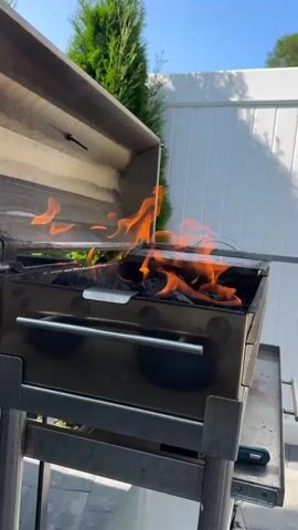 A stainless steel grill with an open lid is positioned outdoors on a paved patio. Flames are actively burning among charcoal briquettes within the grill. A long, slender metal object, possibly a grill tool, is being inserted into the grill from the right side. In the background, a white vinyl fence is visible, with several tall, green evergreen trees situated in front of it. The sky is blue and clear. The scene appears to be in daylight.