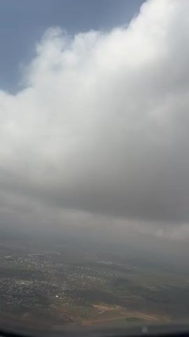 The video shows an aerial view from a plane window.  The sky is mostly covered in thick, grey clouds. Patches of blue sky can be seen through breaks in the clouds. Below the clouds, a landscape of fields and a town can be seen, although visibility is limited due to the cloud cover and haze.