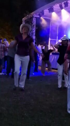 A group of people are dancing in front of a stage with lights. A woman in a cowboy hat and a purple shirt and white pants is in the foreground, dancing. There are other people in the background dancing and socializing. The event appears to be outdoors at night.