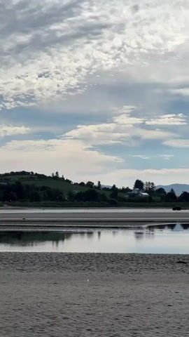 The video shows a tranquil scene of a beach and calm water. In the foreground, there is a sandy beach. Beyond the beach, a body of water reflects the sky and the landscape. The water is shallow in places, revealing sandbars. In the middle ground, across the water, there is a lush green hillside with scattered trees and some houses. Further in the background, there are rolling hills and mountains. The sky is partly cloudy with patches of blue sky visible. There are several small vehicles, possibly ATVs, driving on the sandbars in the water. There are also what appear to be wind turbines visible on the distant hills. The overall impression is one of natural beauty and peacefulness.